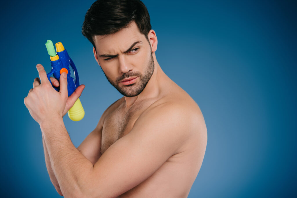 handsome bearded young shirtless man holding water gun and looking away on blue