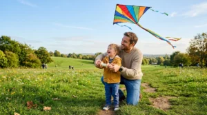 Un père souriant apprend à son enfant à faire voler un cerf-volant dans un parc, illustrant une lettre pour décrire un bon père.